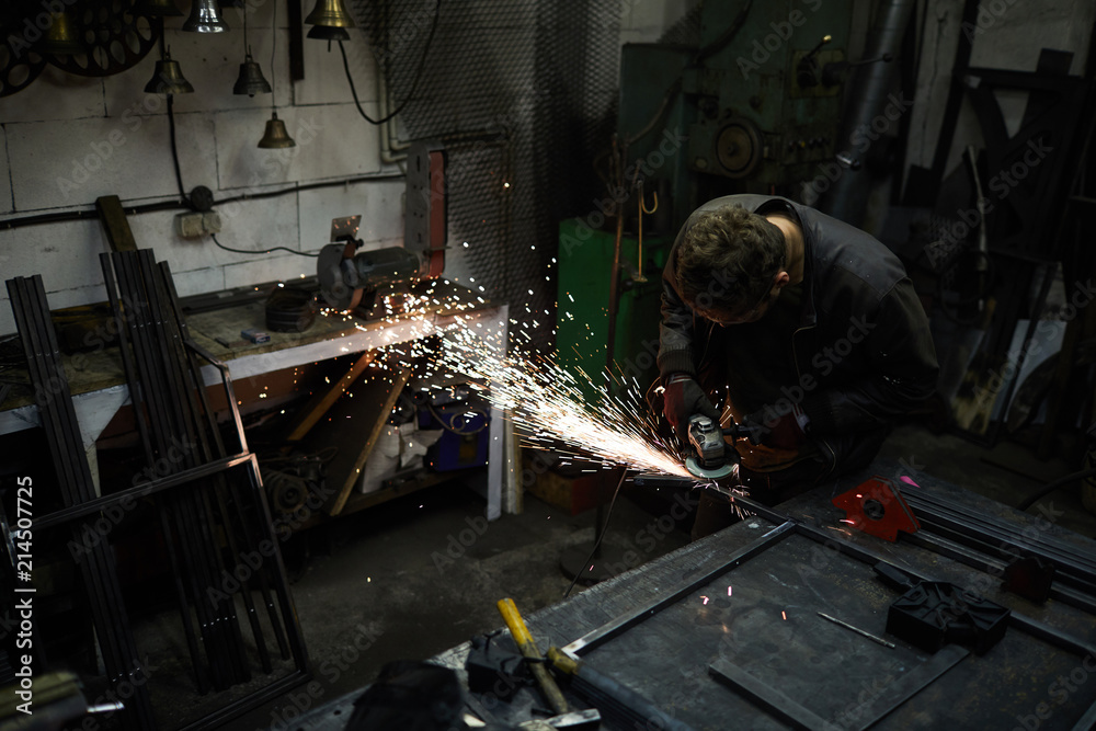 Young man in workwear standing by blacksmith anvil and carrying out sawing work in smithy