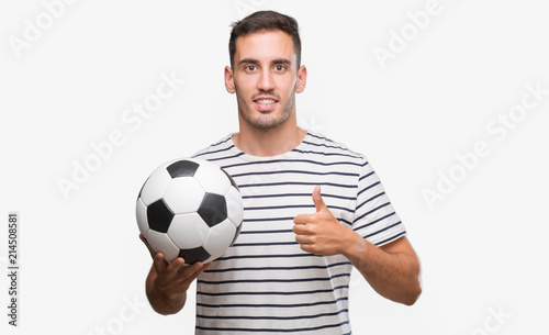 Handsome young man holding soccer football happy with big smile doing ok sign, thumb up with fingers, excellent sign © Krakenimages.com