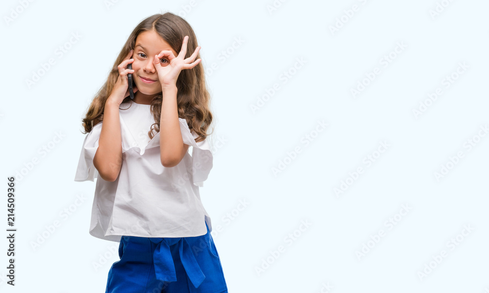 Brunette hispanic girl using smartphone with happy face smiling doing ok sign with hand on eye looking through fingers Brunette hispanic girl using smartphone with happy face smiling doing ok sign with hand on eye looking through fingers