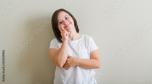 Down syndrome woman standing over wall looking confident at the camera with smile with crossed arms and hand raised on chin. Thinking positive.
