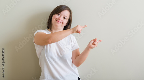 Down syndrome woman standing over wall smiling and looking at the camera pointing with two hands and fingers to the side. photo