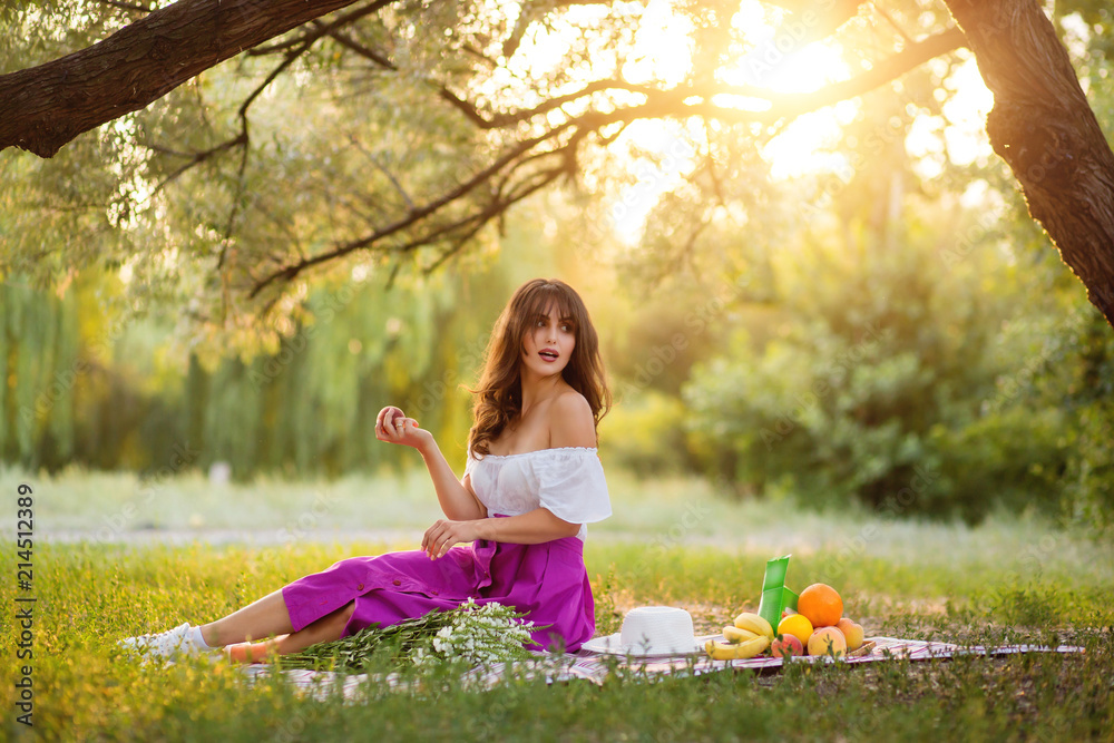 Fototapeta premium Beautiful girl with a peach in her hands on a picnic in the park. Looking away