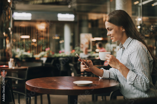 Nice and young girl is sitting at table and looking at phone. She is holding it and smiling. Also girl has a cup of coffee in other hand. She has lunch. © Anton