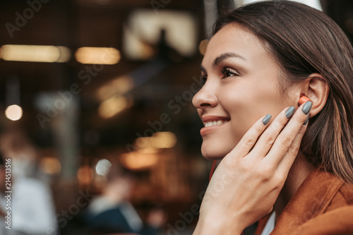 Relaxed and chilled girl is looking forward and smiling. She is holding hand on ear. Woman with brown hair looks happy