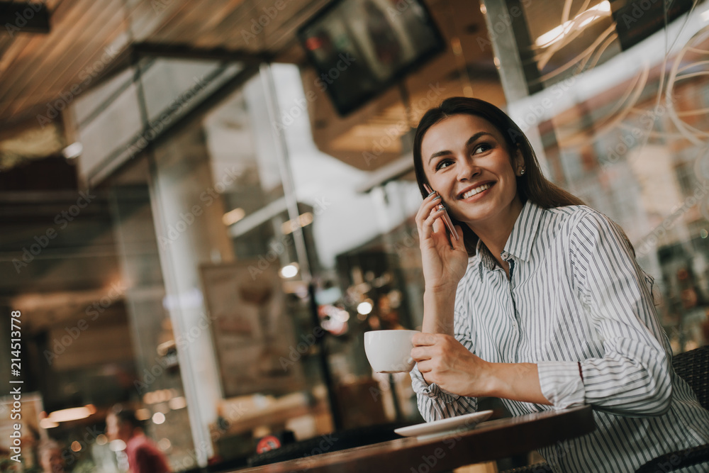 Portrait of business woman sitting at table in cafe. She is talking on the phone and smiling. Girl is looking to the side. She is holding cup of coffee in roght hand. Woman looks happy