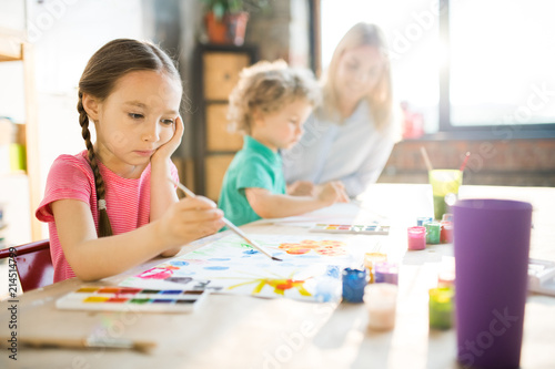 Concentrated little girl sitting at the table and painting picture with young woman and boy in the background