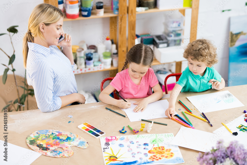 Fototapeta premium Children sitting at wooden table and drawing a picture with colorful pencils with teacher talking on cellphone and looking at how her pupils work