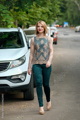 The blond girl in a fashionable green overalls goes along the asphalt road near to the cars.