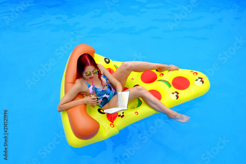 A young woman wearing a bathing suit lies relaxed on an  inflatable swim bed. She takes time out to read a book while she enjoys the pool atmosphere.