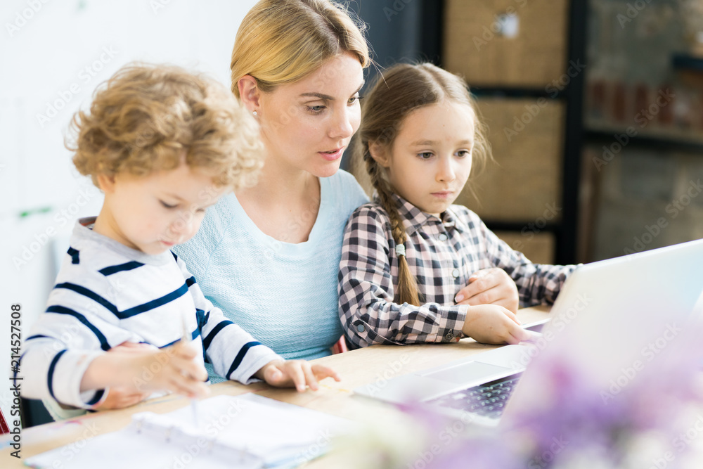 Beautiful mother sitting at the table together with her little daughter and son and looking at laptop Beautiful mother sitting at the table together with her little daughter and son and looking at laptop