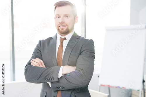 Waist up portrait of successful young businessman posing confidently standing with arms crossed in modern office