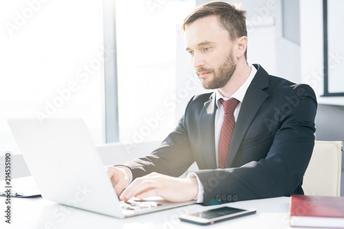 Portrait of successful businessman using laptop while working at desk in office, typing, copy space