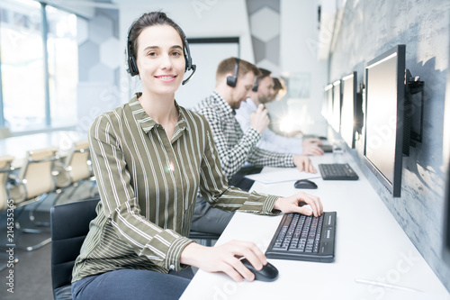 Portrait of smiling young woman wearing headset looking at camera while working with group of help desk operators sitting in row, copy space