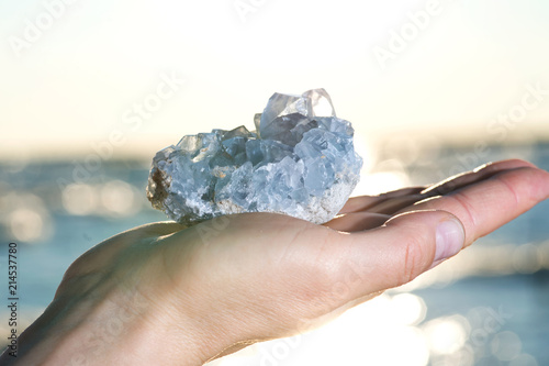 Принти на полотні Blue Celestite cluster from Madagascar in woman's hand at sunrise in front of  the lake