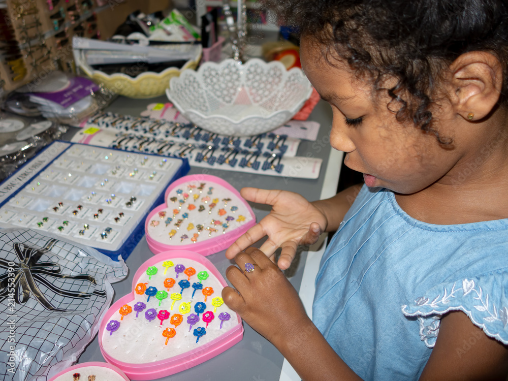 Mixed ethnicity little girl choosing a ringlet in the shop. Baby-girl trying the ring on the finger of hand Mixed ethnicity little girl choosing a ringlet in the shop. Baby-girl trying the ring on the finger of hand