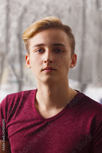 Handsome blond boy teenager in t-shirt poses near window in room