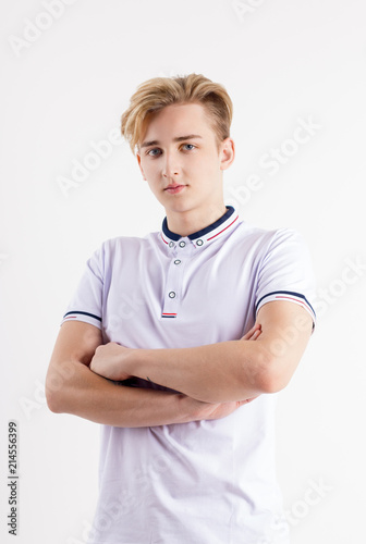 Handsome boy teenager in white shirt stands with crossed arms in white studio