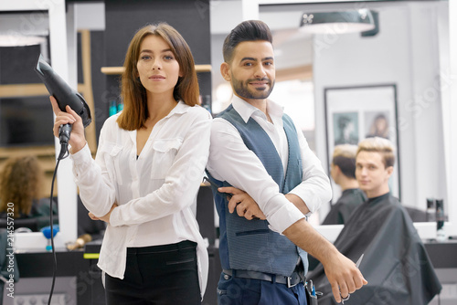 Two hairstylers posing standing in modern beaty salon, young client sitting behind. Hairdressers wearing classic white shirts, man having grey waistcoat. Woman keeping hair dryer, looking at camera. photo