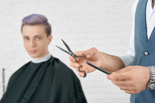 Barber's hands holding metallic scissors and black plastic comb. Young client with toned lilac hair sitting behind and looking at camera. Soft focus. Posing on white brick wall background.
