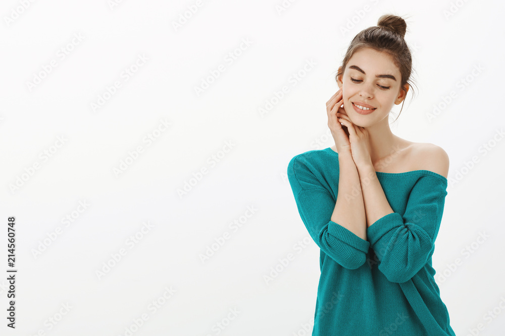 Feminine sensual and tender female with bun hairstyle in stylish loose sweater, gently touching face and smiling while gazing down with cute expression, enjoying beautiful and clean face Feminine sensual and tender female with bun hairstyle in stylish loose sweater, gently touching face and smiling while gazing down with cute expression, enjoying beautiful and clean face