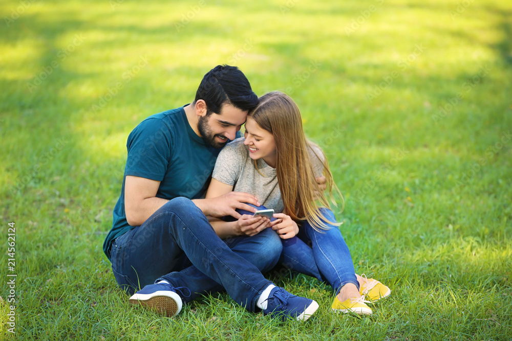 Happy young couple with mobile phone sitting on green grass in park Happy young couple with mobile phone sitting on green grass in park