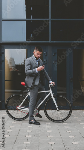 Handsome young adult man wearing suit checking phone before riding his classic bicycle to work in the morning