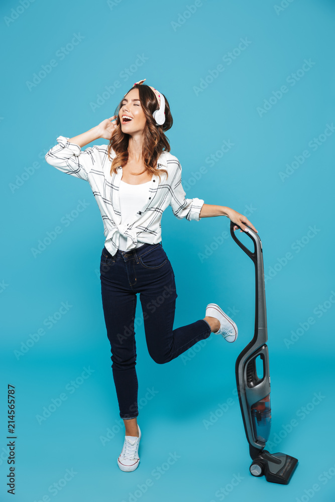 Full length portrait of joyful young woman 20s listening to music via wireless headphones while housekeeping and vacuuming floor, isolated over blue background Full length portrait of joyful young woman 20s listening to music via wireless headphones while housekeeping and vacuuming floor, isolated over blue background