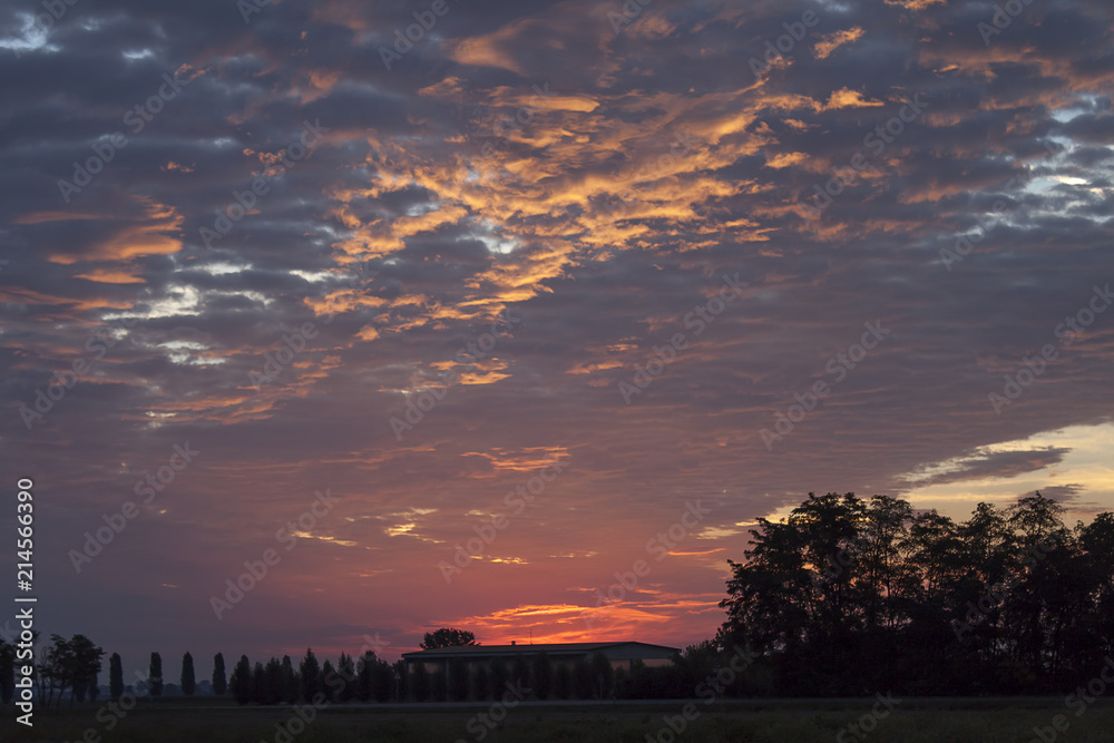 Obraz premium Over the fields and trees a beautiful sunset. Rays of the sun through multicolored cumulus clouds