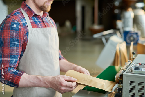 Mid section of unrecognizable man wearing apron packing freshly roasted coffee beans in craft paper bags while working in artisan roastery house, copy space
