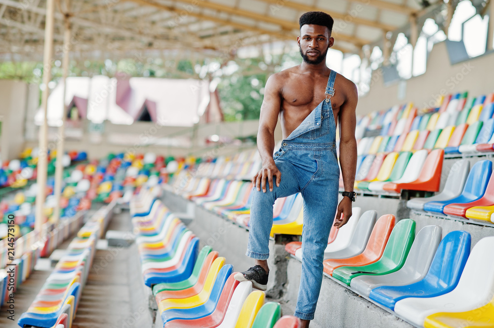Handsome sexy african american bare torso man at jeans overalls posed on colored chairs at stadium. Fashionable black man portrait. Handsome sexy african american bare torso man at jeans overalls posed on colored chairs at stadium. Fashionable black man portrait.