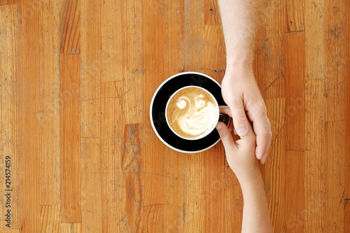 Young couple in love holding hands over black cup of coffee with swan shaped foam latte art on vintage grunged scratched table. Man & woman body language. Background, top view, close up, copy space.