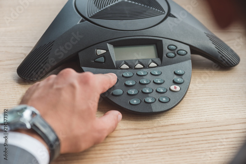 cropped shot of businessman with stylish wristwatch pushing button of conference phone on table at office