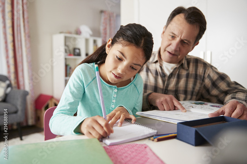 Grandfather Helping Granddaughter With Homework Sitting At Desk In Bedroom