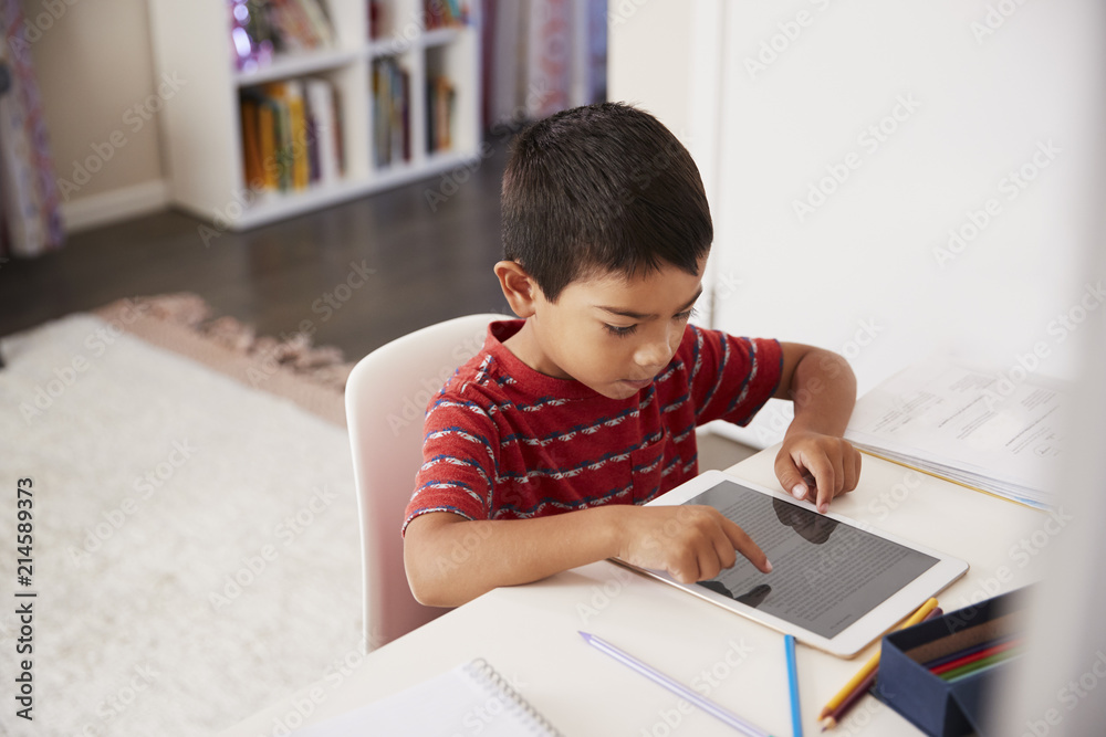 Young Boy Sitting At Desk In Bedroom Using Digital Tablet To Do Homework Young Boy Sitting At Desk In Bedroom Using Digital Tablet To Do Homework