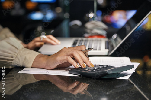 Crop side view of female hands in light blouse sitting at dark desk with laptop and papers and using calculator while counting income on blurred background.