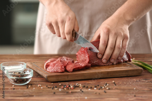 Obraz na plátně Man cutting raw meat on wooden board in kitchen