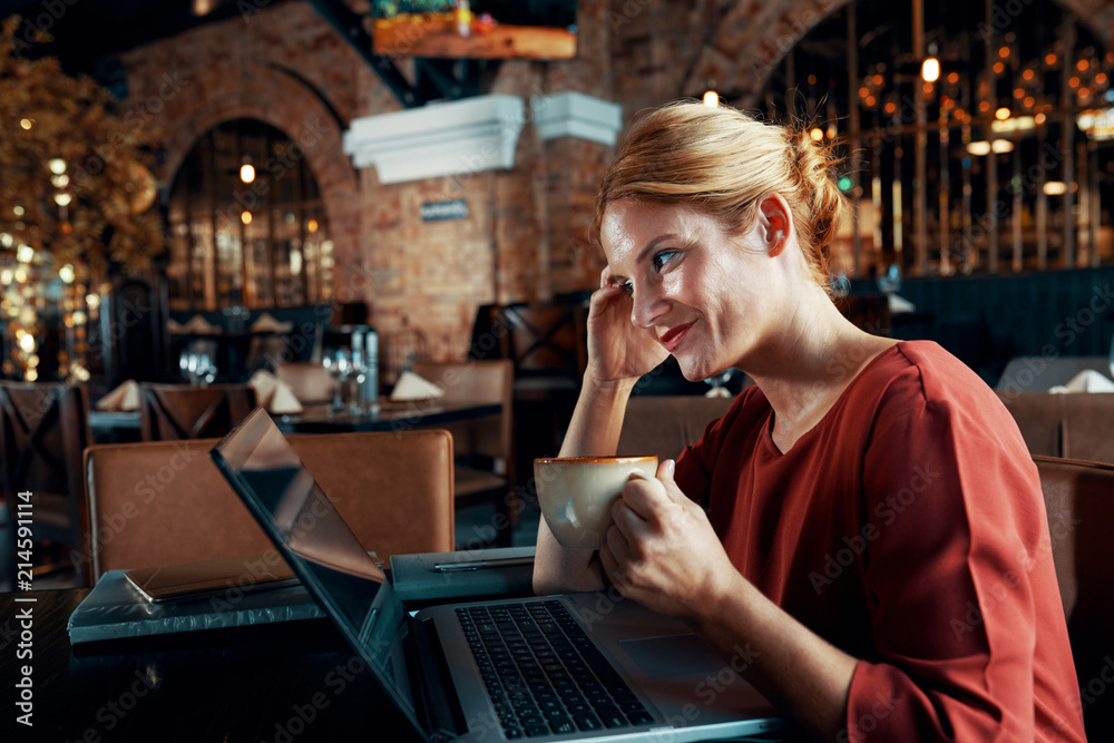 Side view of pretty young woman using wi-fi free and communicating on laptop while drinking coffee at the restaurant
