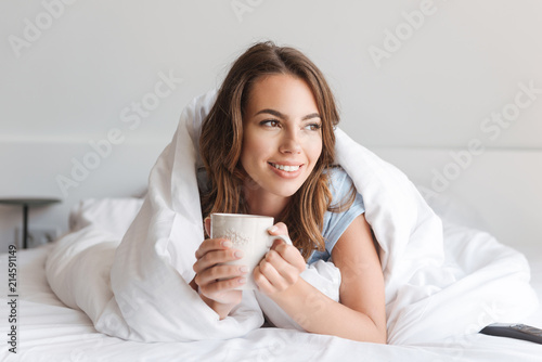 Smiling young woman holding mug while laying in bed