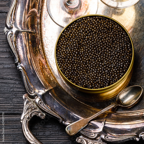 Black caviar in can and champagne on silver tray on black wooden background close-up