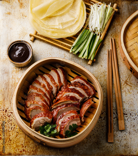 Sliced Peking Duck in bamboo steamer served with fresh cucumber, onion, Hoysin sauce and roasted wheaten pancakes on metal background