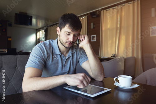 Young male blogger working on touchpad using wireless internet connection while sitting at wooden table in cozy coffee shop. Freelancer using contact information of job vacancy calling on mobile phone photo