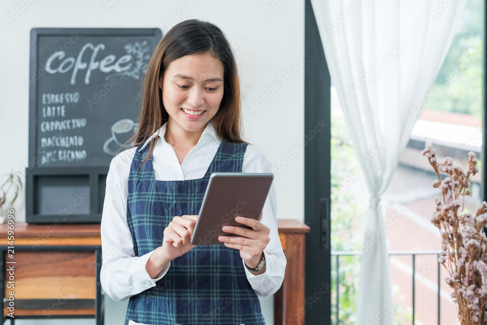 Fototapeta premium Asian female barista wear blue apron hold tablet computer coffee menu with smiling face in cafe waiting order from customer ,Coffee business owner concept.