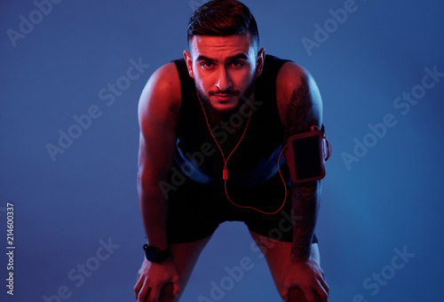 Serious adult athletic man in headphones and armband leaning on knees looking at camera with determination