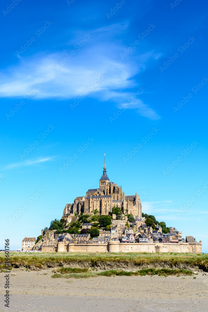 General view of the Mont Saint-Michel tidal island, located in France on the limit between Normandy and Brittany, from the bay at low tide under a summer blue sky with salt meadow in the foreground.
