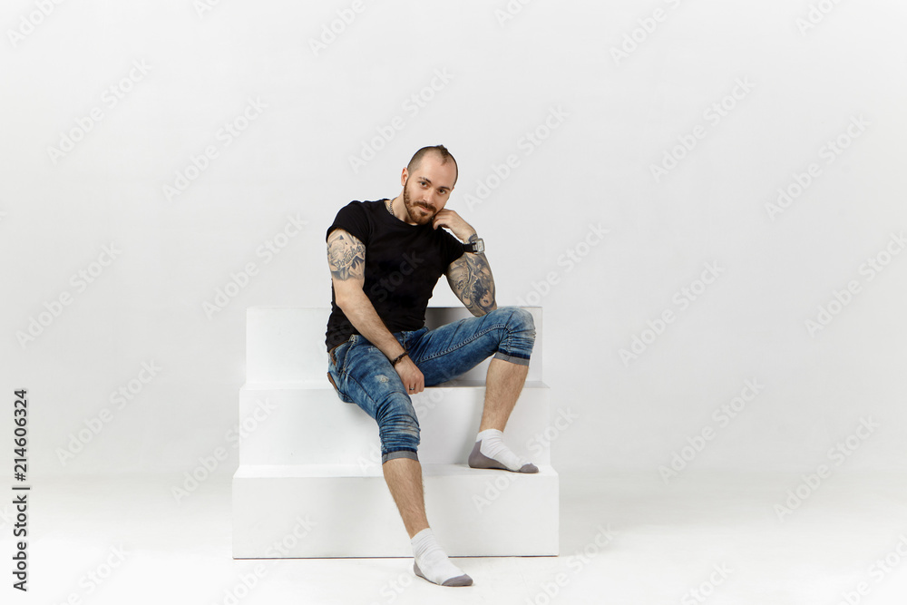 Trendy looking hipster guy with stubble and tattooed body having relaxed look, sitting on white stairs against blank studio wall background with copyspace for your text or advertising content Trendy looking hipster guy with stubble and tattooed body having relaxed look, sitting on white stairs against blank studio wall background with copyspace for your text or advertising content