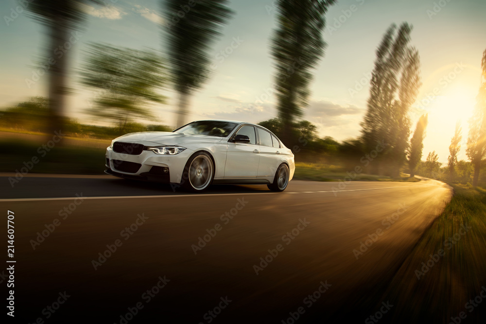 White car is driving on empty countryside asphalt road at sunset - obrazy, fototapety, plakaty 