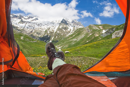 View from orange tent with feet of resting tourist or hicker on snowy mountain peaks and green valleys on sunny day. Alps, France. Lifestyle concept