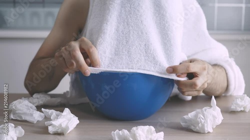 Sick young man with the flu doing some inhalations at home from a bowl. He covers his head and put his face right above the bowl that has steam coming out of it.