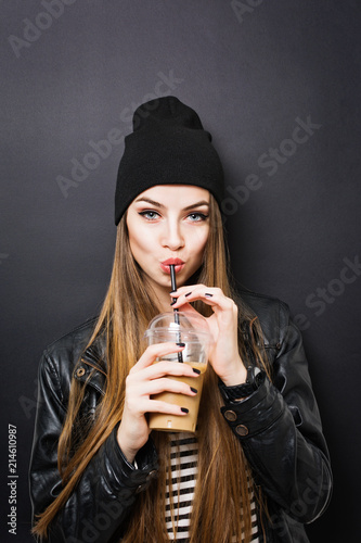Teenage girl drinking takeaway coffee from a straw, wearing black outfit, against black background