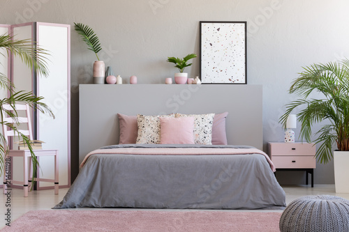 Poster on grey bedhead in bedroom interior with pink pillows on bed next to chair. Real photo photo